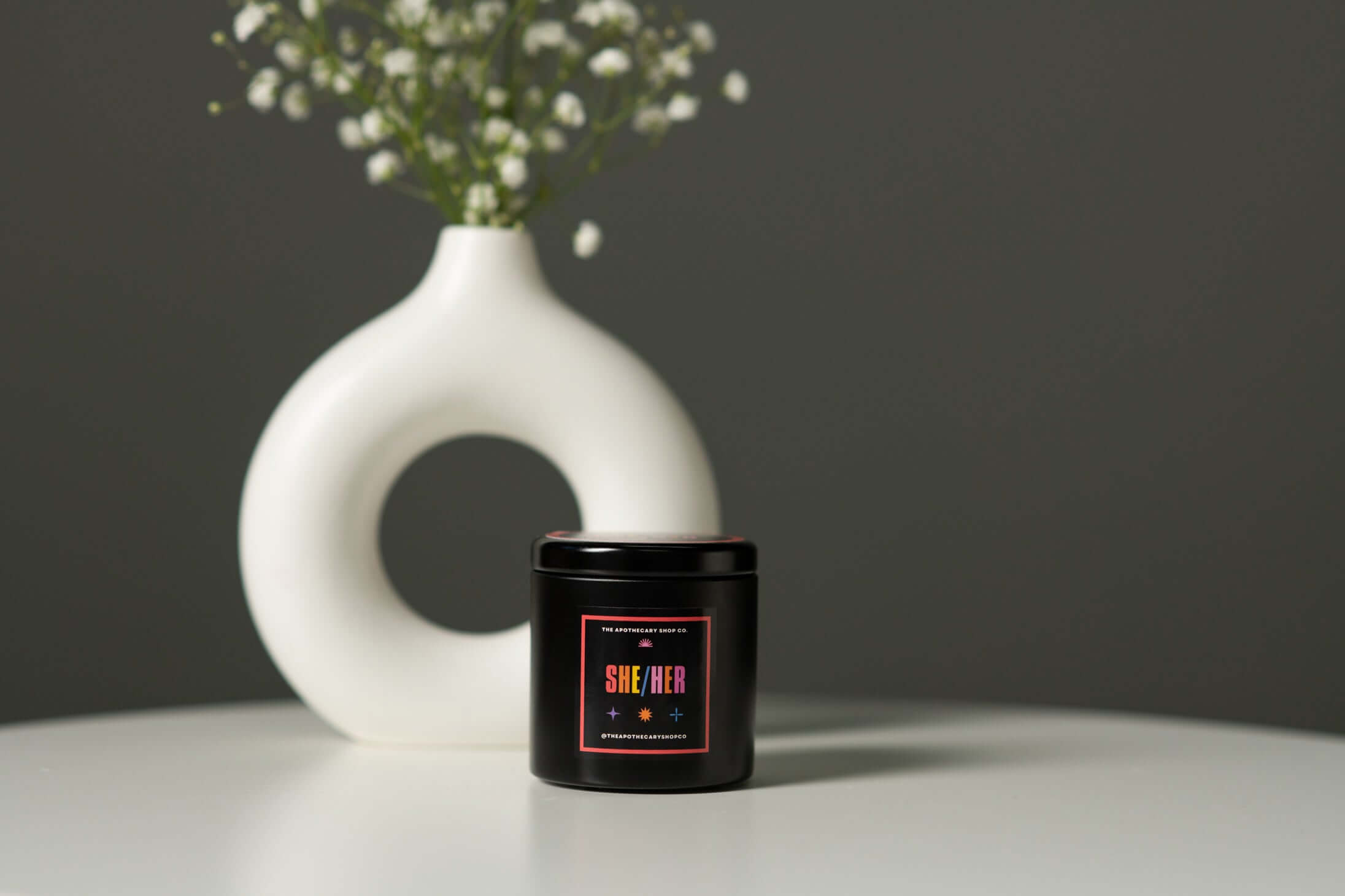 Gender Fluid Candle - She/her 4 Oz Ocean Tide in a black metal tin next to a stylish white vase with flowers.