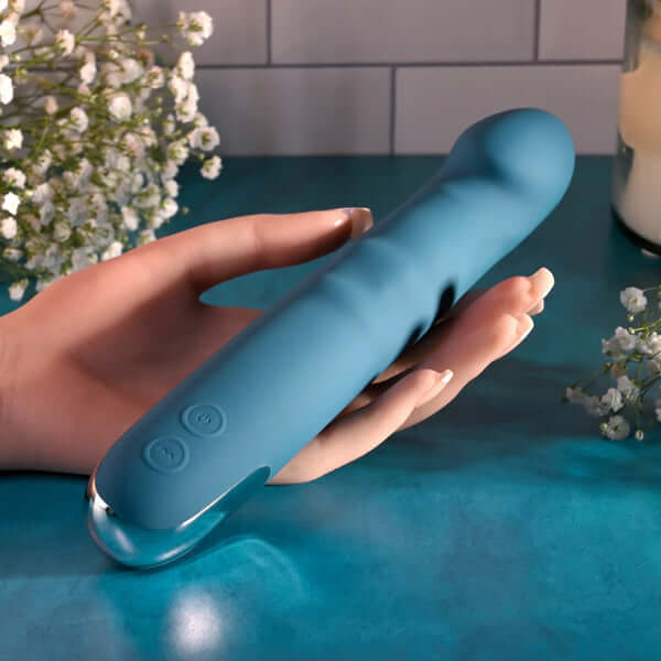 Queen of All Trades blue silicone dual vibrator held in a hand, surrounded by flowers on a teal surface.