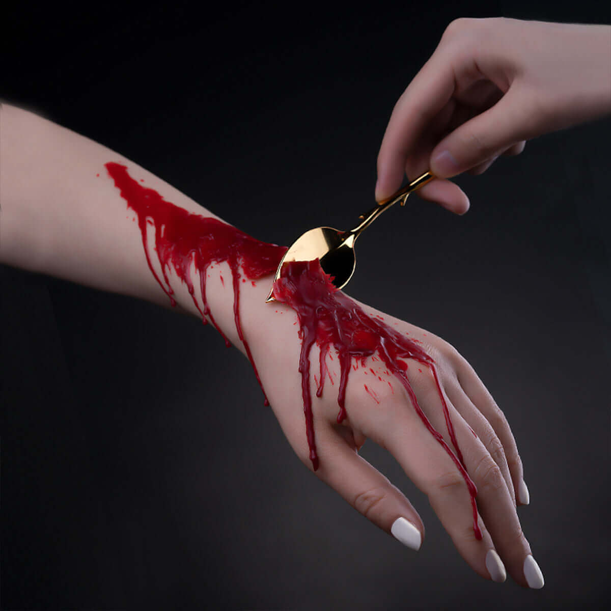 Hand using a gold spoon to apply red liquid on another hand, creating a dramatic artistic effect.