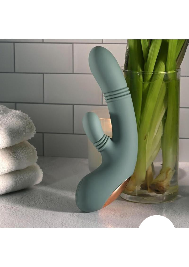Rub Me Tender Thrusting Vibrator with Clitoral Stimulation Green placed beside a glass vase with fresh flowers.