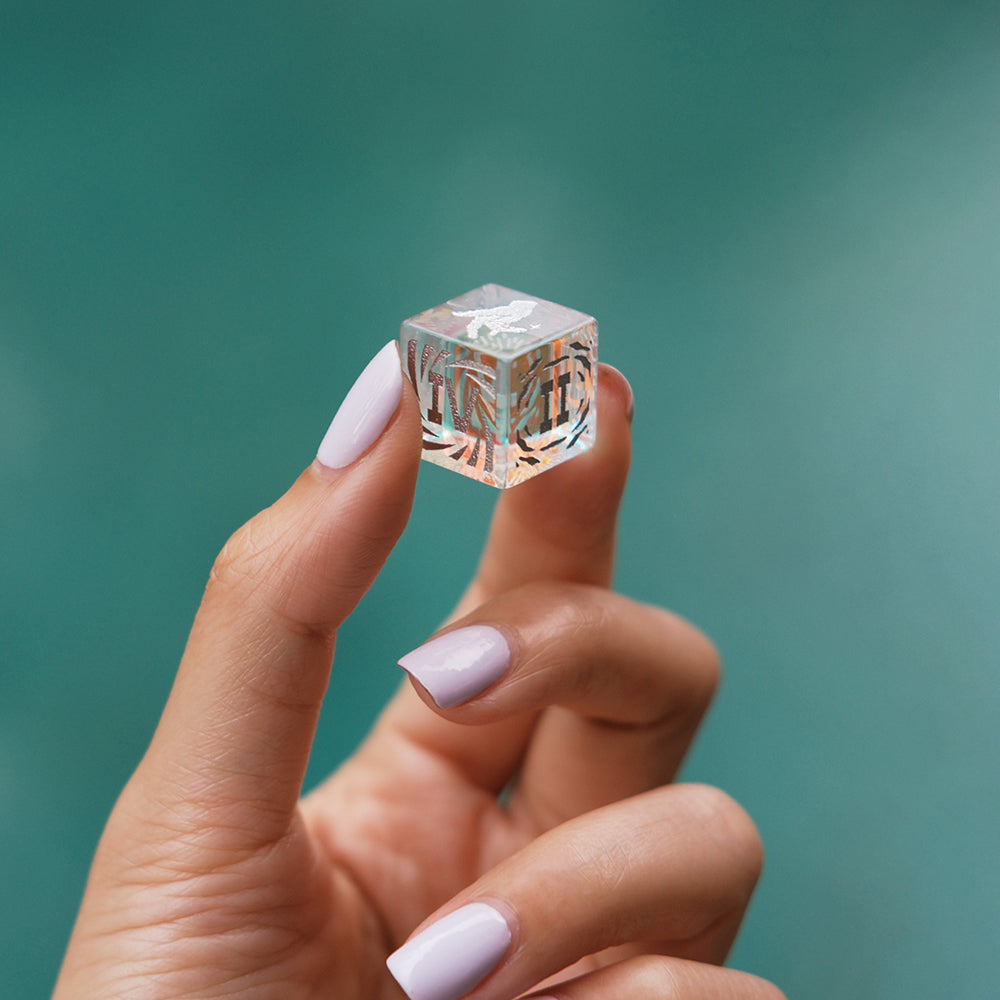 A hand holding a small clear cube with intricate designs, perfect for enhancing gameplay in Biird X Jouissance Club Together Couples Card Game.