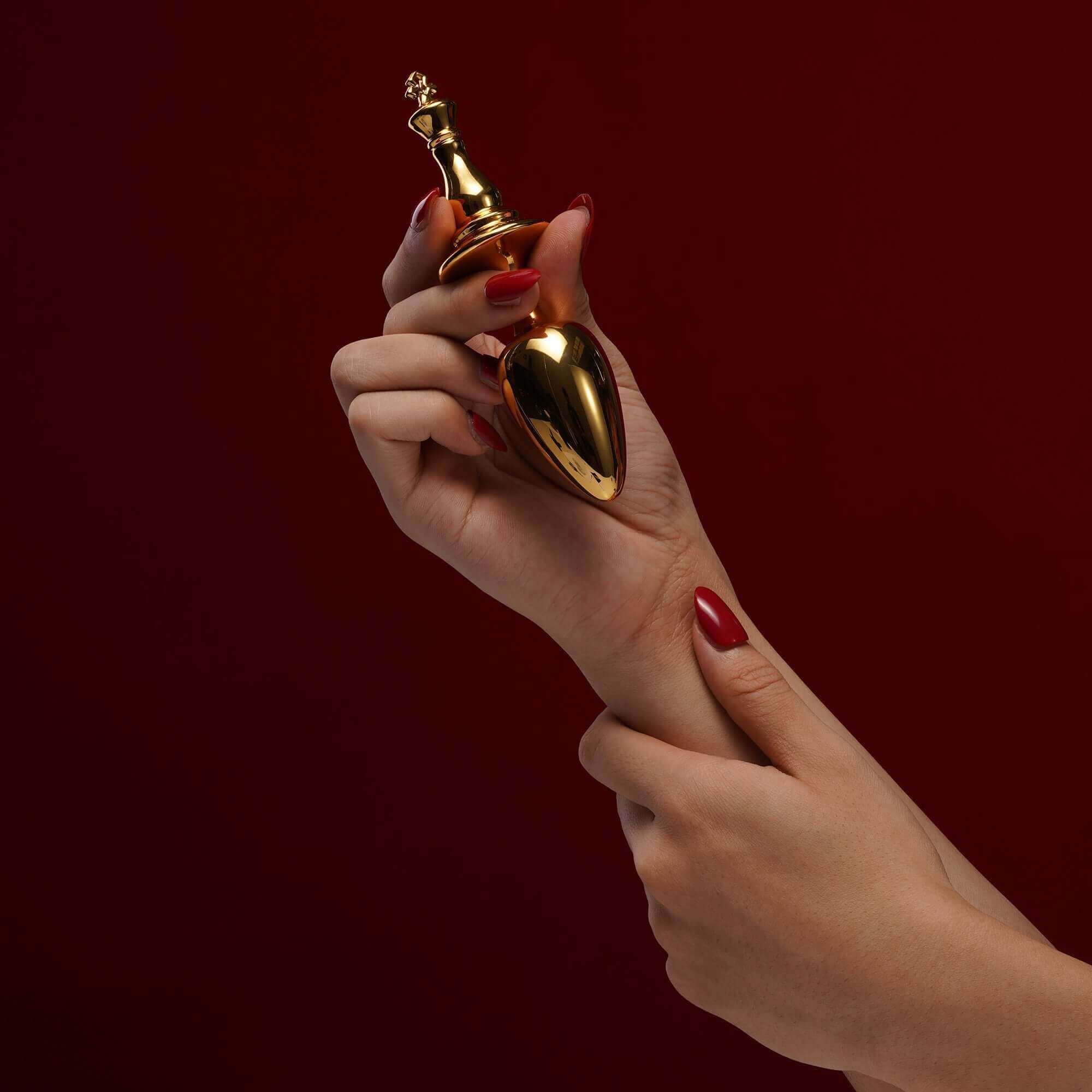 Hand holding the King Butt Plug from "The Chess" Collection, showcasing its gold electroplated design against a dark background.