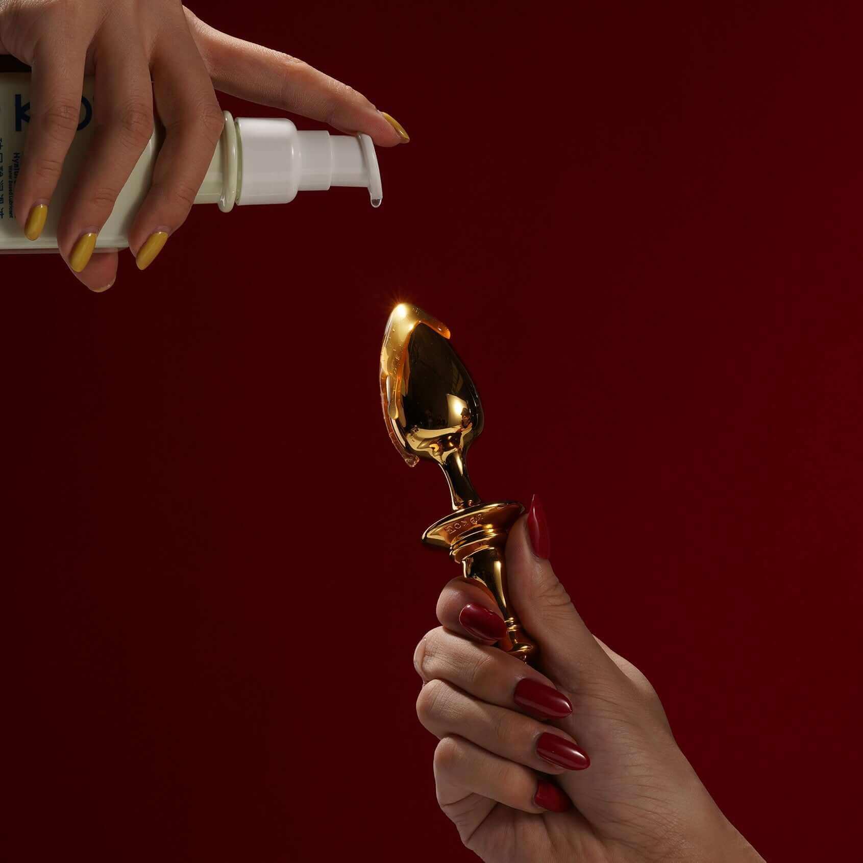 Person holding a gold King butt plug while another hand sprays lubricant, set against a deep red background.