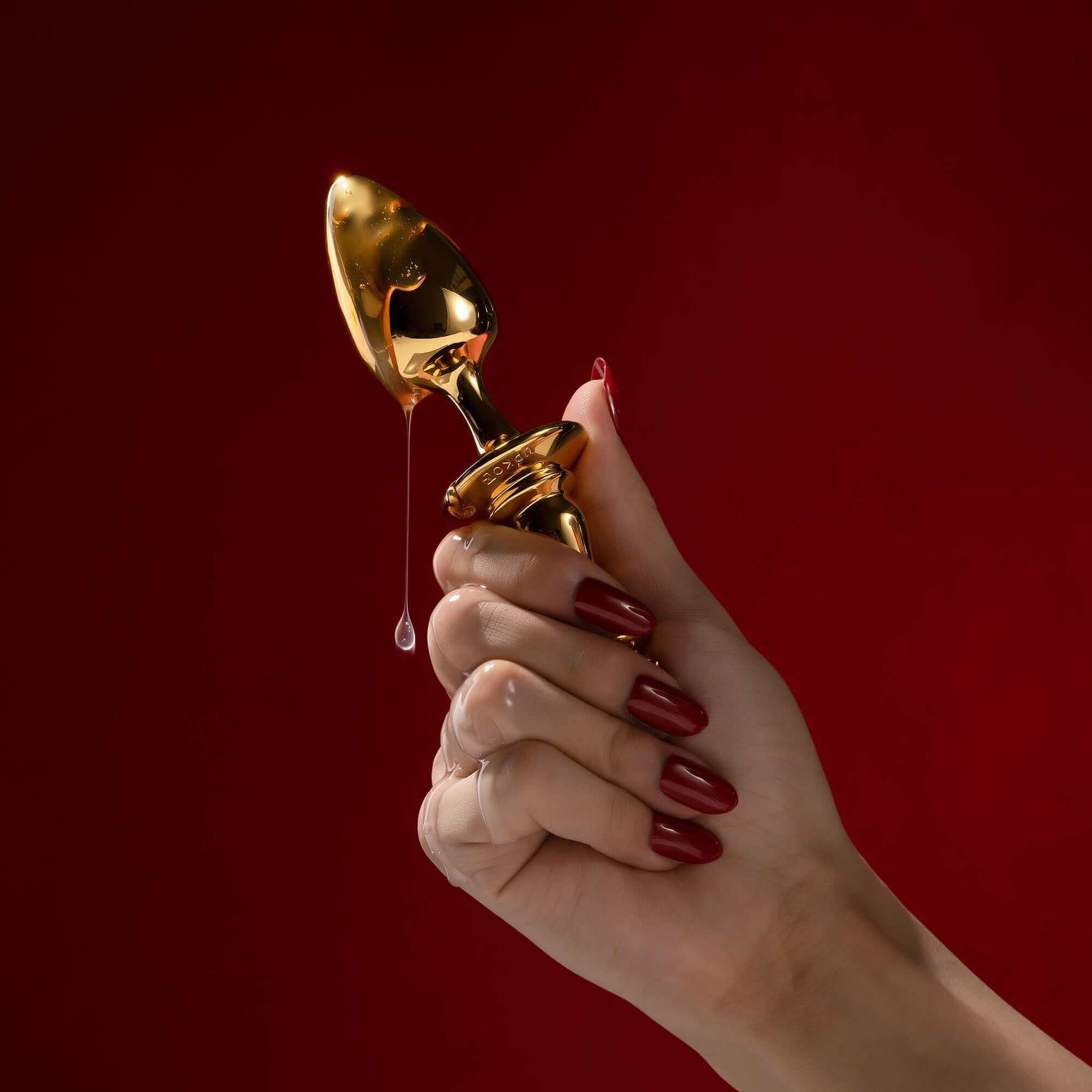 Hand holding golden King Butt Plug from "The Chess" Collection against a red background, showcasing its sleek design.