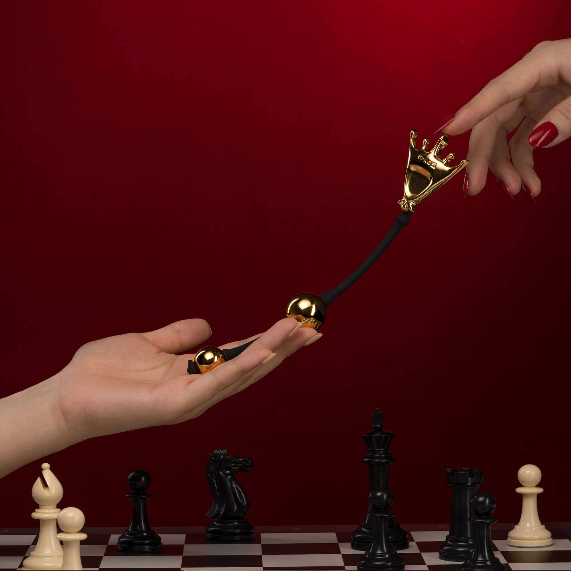 Queen Premium Metal Pleasure Balls from The Chess Collection being held against a chessboard backdrop.