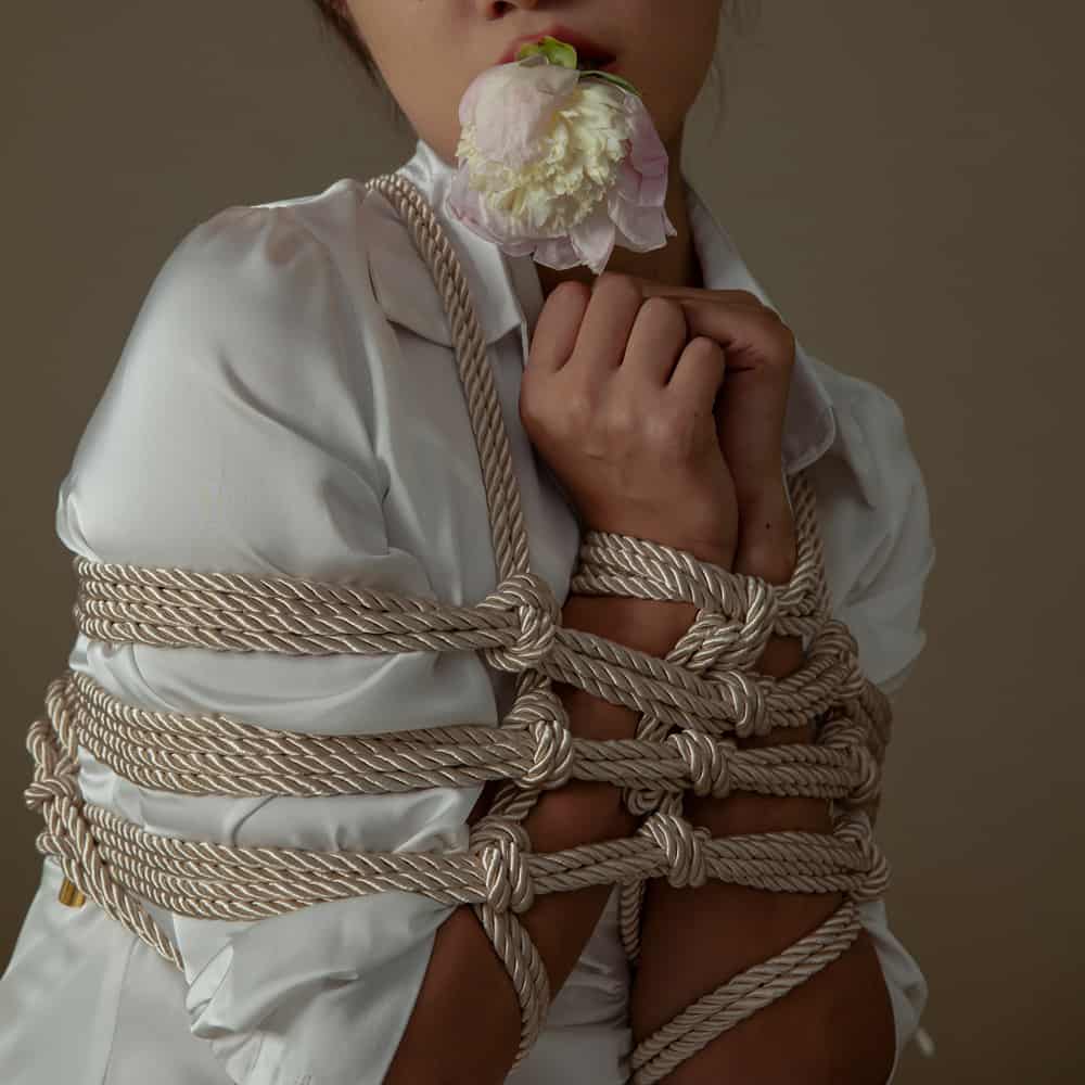 Model demonstrating Shibari bondage technique using luxurious rope with a flower in her mouth.