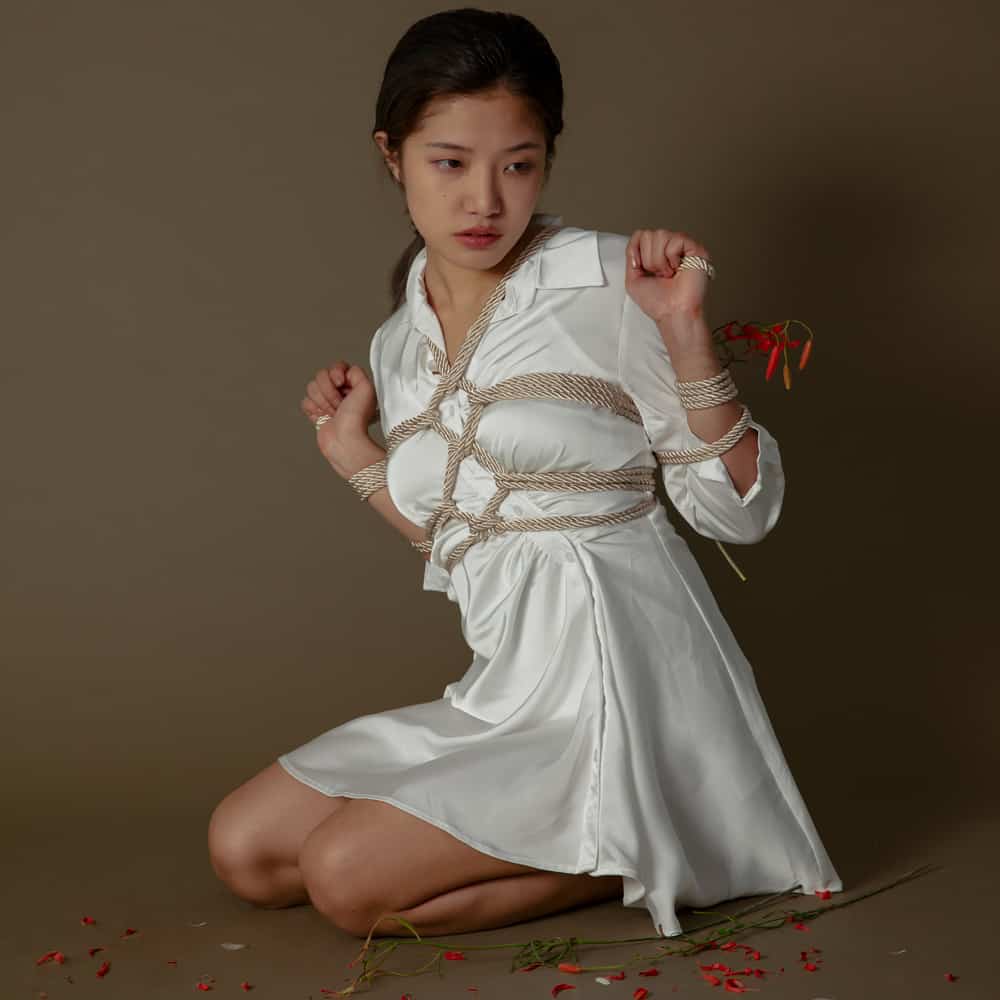 Model wearing white dress tied with luxurious shibari rope, demonstrating bondage techniques in a styled setting.